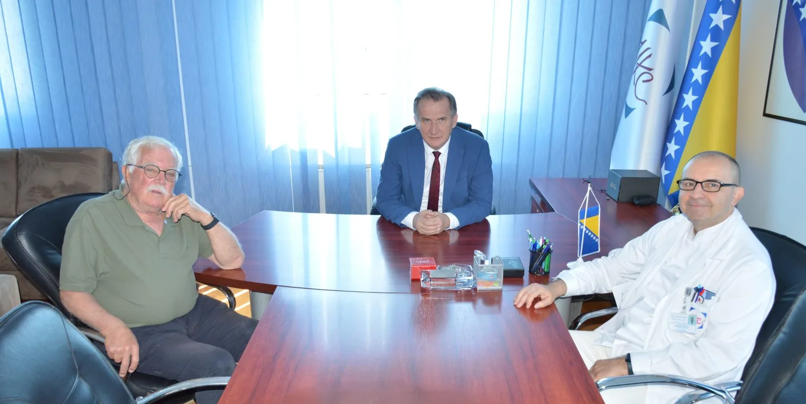 Tri muškarca sjede za stolom u uredskom prostoru ispred zastava Bosne i Hercegovine i drugih simbola.