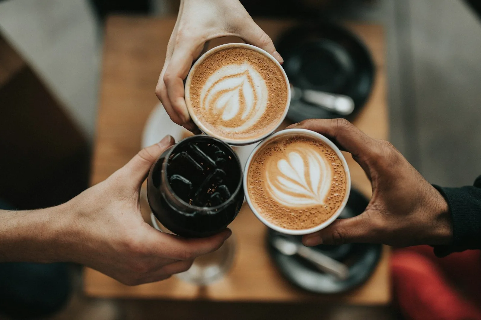 Tri ruke drže šoljice kafe, jedna sa ledenom kafom, a dvije sa latte artom.