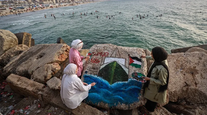 Tri žene u maramama farbaju veliki kamen uz obalu mora, oslikavajući brod i palestinsku zastavu.