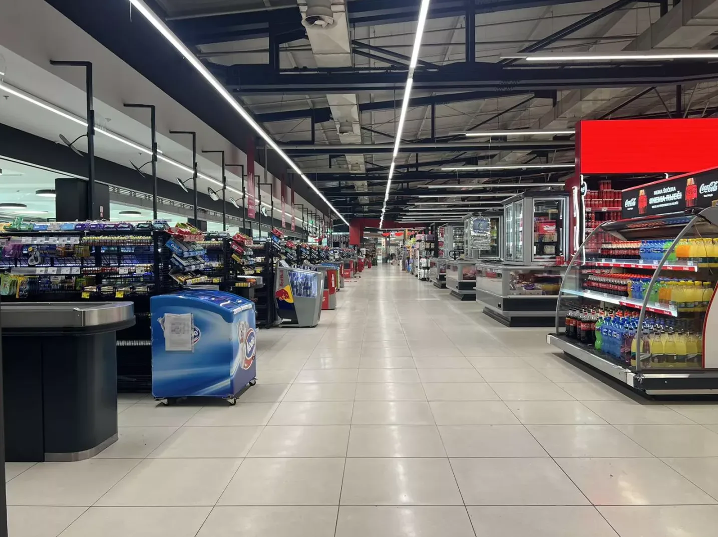 Unutrašnjost modernog supermarketa s brojnim rafaovima punim proizvoda, rashladnim vitrinama s pićima i aparatima za sladoled, osvijetljena dugim nizovima LED rasvjete na plafonu.