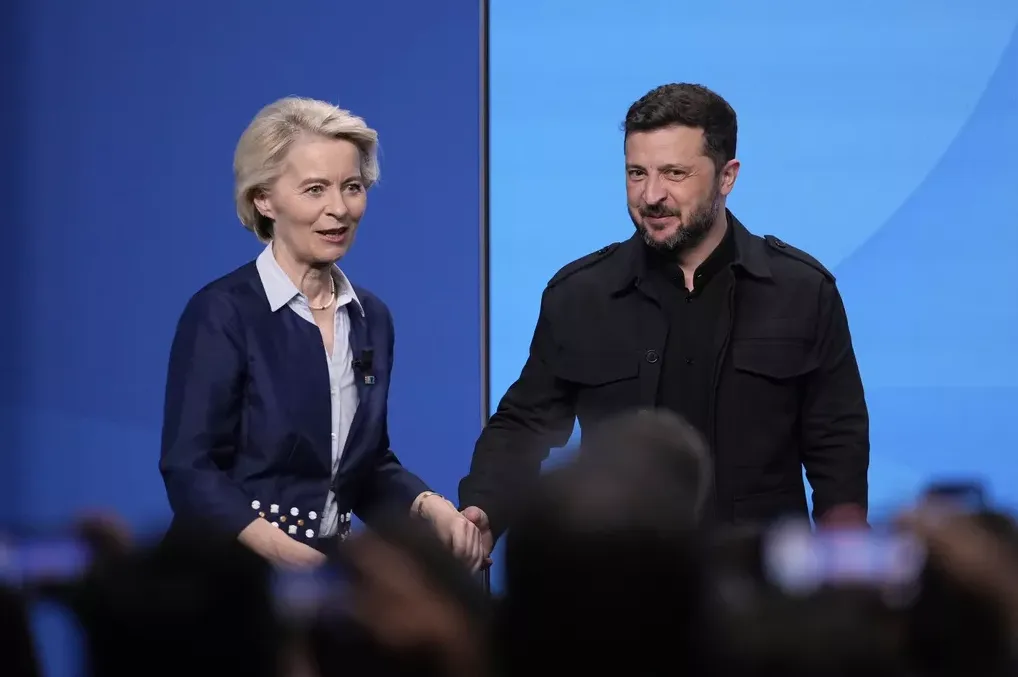 Ursula von der Leyen i Volodimir Zelenski se rukuju ispred plave pozadine.