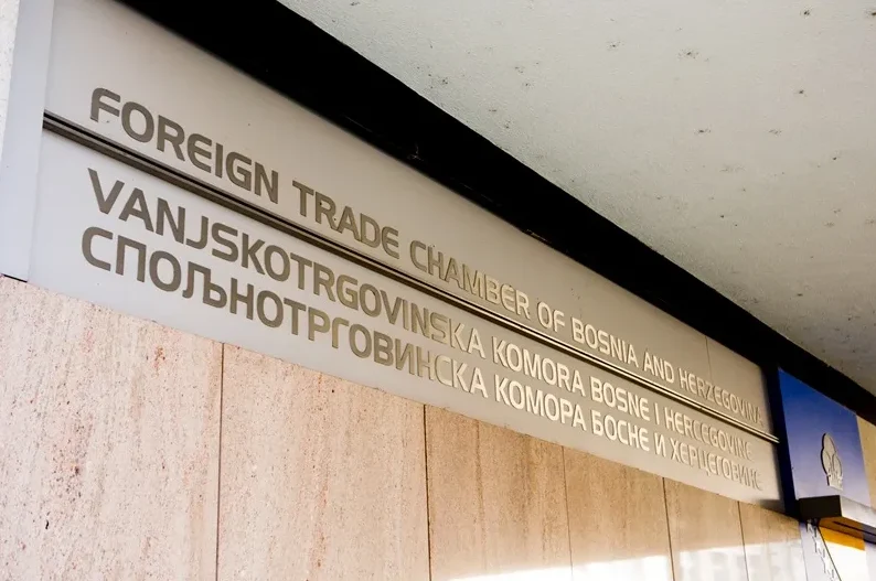 Natpis na tri jezika: 'Foreign Trade Chamber of Bosnia and Herzegovina', 'Vanjskotrgivinska komora Bosne i Hercegovine' i 'Внешнеторговая палата Боснии и Герцеговины', ispisan na bijeloj ploči iznad mermerne fasade.