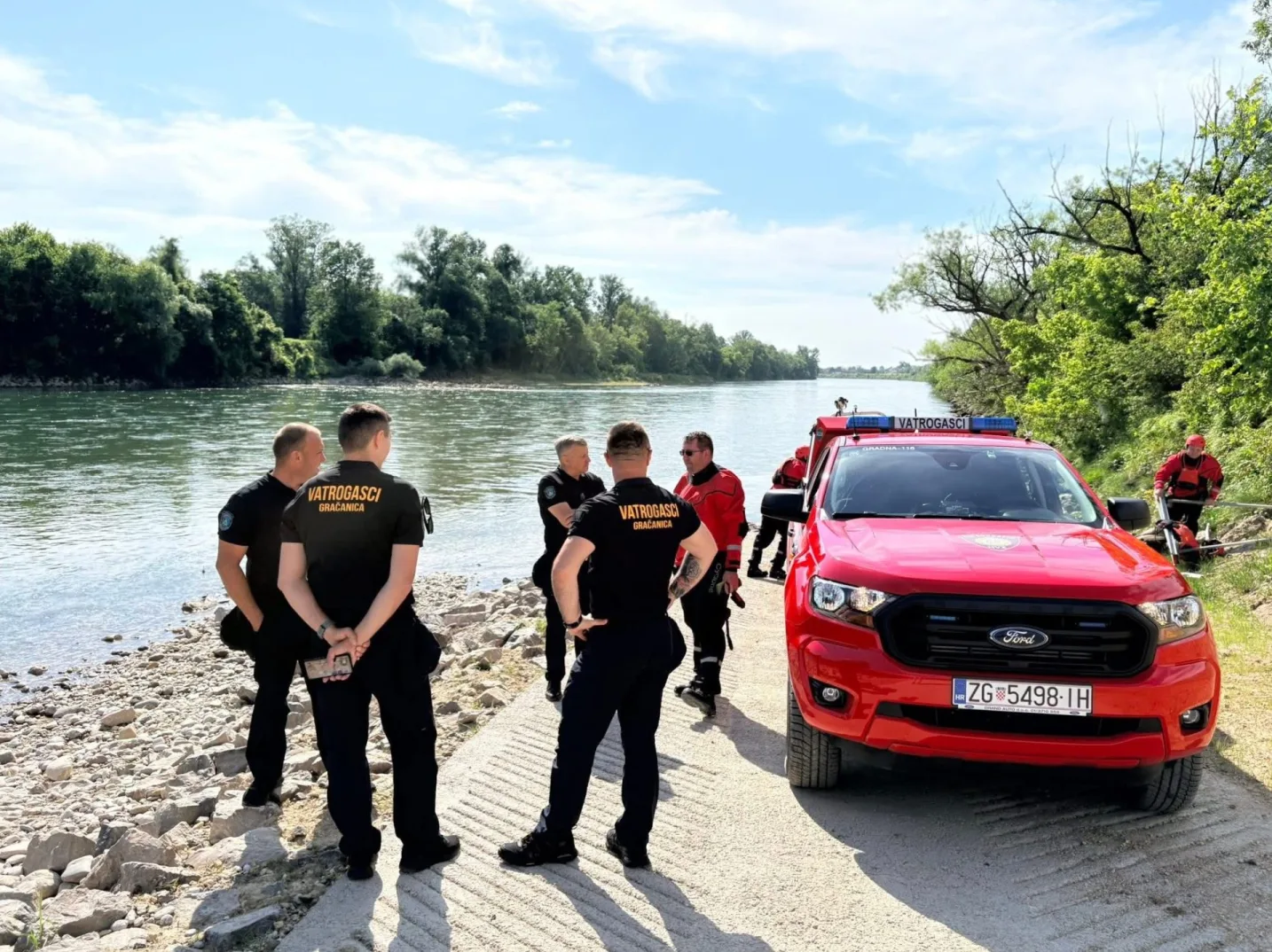 Vatrogasci u crnim uniformama i crvenim jaknama stoje na obali rijeke pored crvenog Ford Ranger vozila.