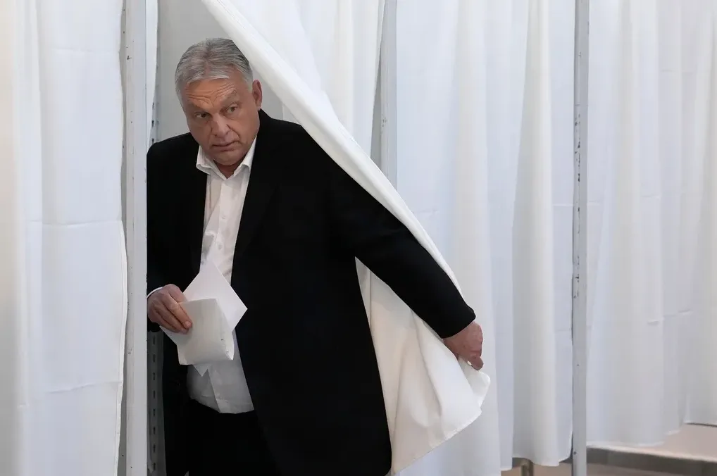 Viktor Orban u crnom odijelu i bijeloj košulji izlazi iza bijele zavjese u kabini za glasanje, držeći papire u ruci.