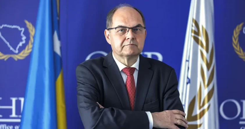 Portret muškarca u tamnom odijelu, bijeloj košulji i crvenoj kravati sa bijelim šarama, sa naočarima i prekriženim rukama.