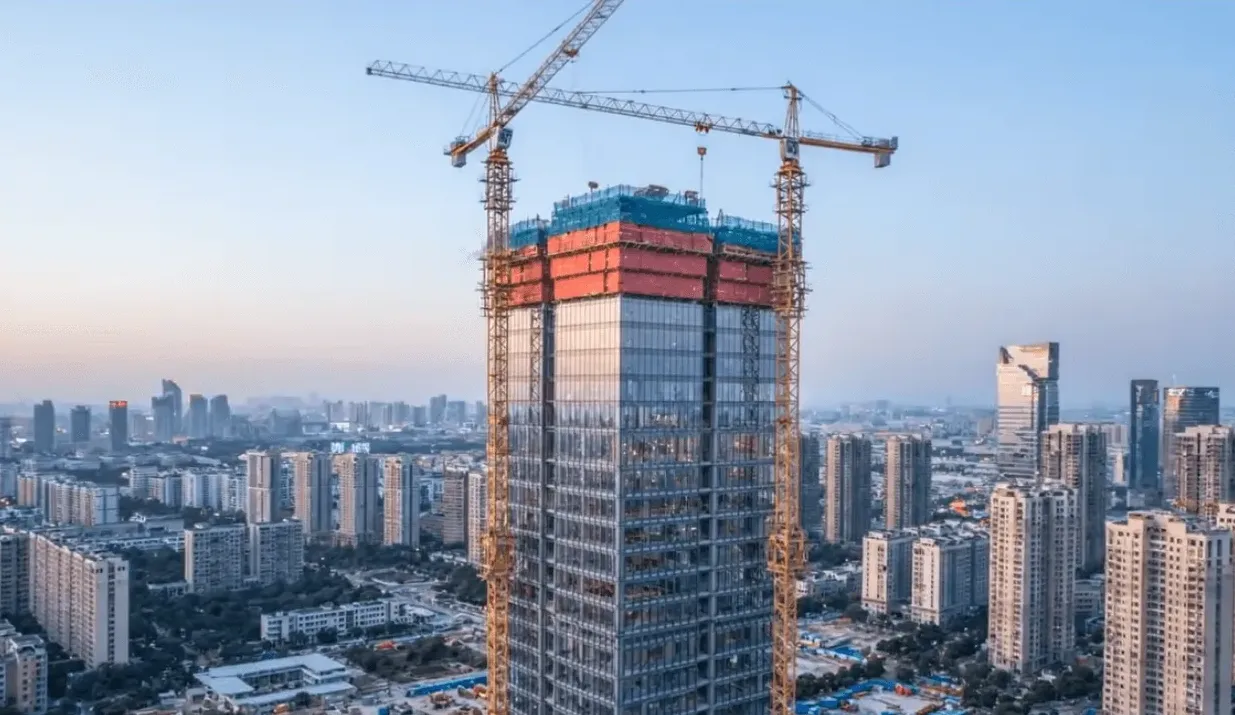 Fotografija visokog solitera u fazi izgradnje, sa dva velika građevinska krana iznad vrha zgrade.