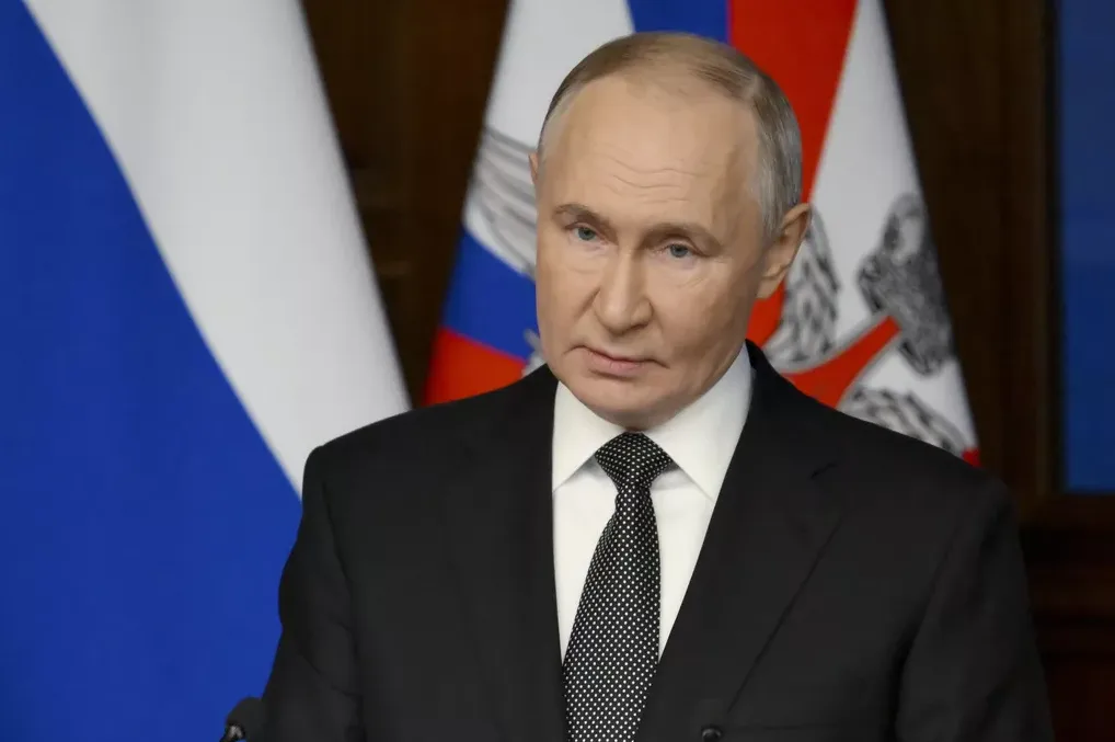 Vladimir Putin u crnom odijelu i bijeloj košulji s crnom tačkastim kravatom, govori pred ruskim zastavama.