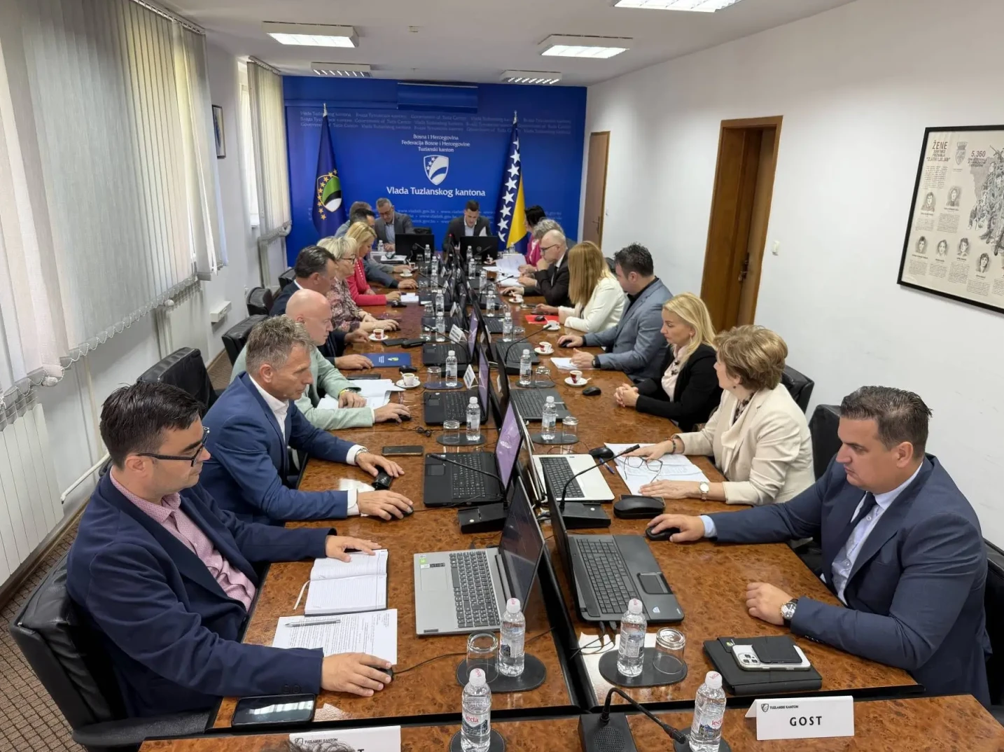 Grupa ljudi sjedi za velikim drvenim stolom u sali za sastanke, ispred svakog od njih nalazi se laptop, papiri i flašice vode. Na zidu iza njih nalaze se zastave BiH i Federacije BiH, te grb Tuzlanskog kantona. Na stolu su primjetne oznake sa imenima, jedna od njih glasi "GOST".
