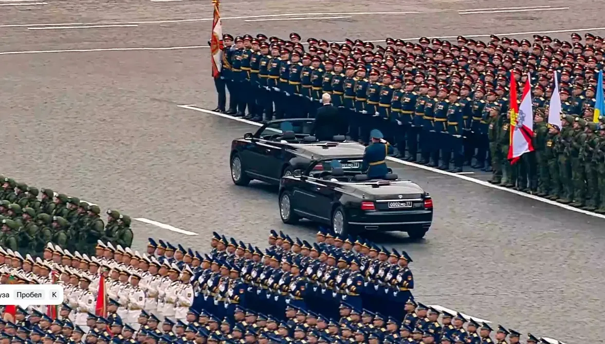 Dva crna kabrioleta voze pored postrojenih vojnika u uniformama, dok visoki zvaničnici stoje u vozilima i salutiraju.