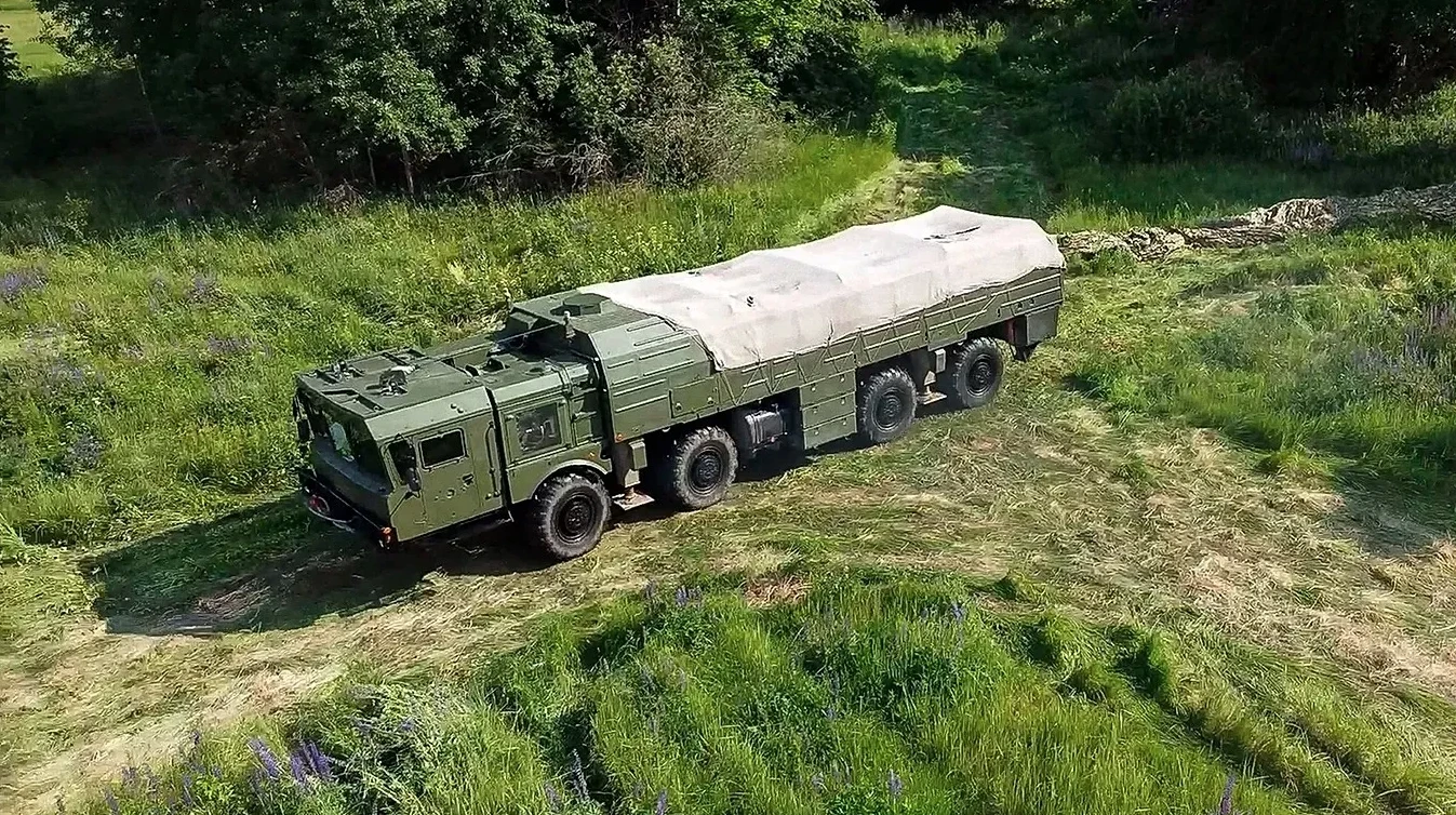 Zeleni vojni kamion sa više osovina prekriven ceradom vozi po neravnom terenu obraslom visokom travom i cvijećem.