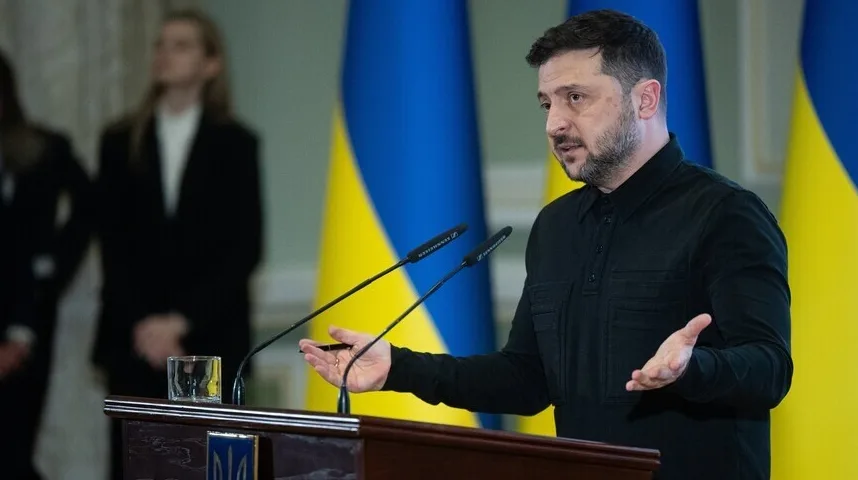 Volodimir Zelenski govori pred mikrofonom iza podijuma, ukrajinska zastava u pozadini