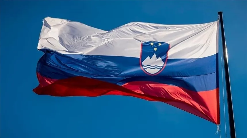 Zastava Slovenije sa grbom koji prikazuje planinu Triglav i dvije zvijezde, vijori se na jarbolu pod vedrim plavim nebom.
