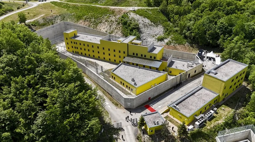 Moderan zatvor žute boje sa betonskim zidovima okružen zelenilom i šumom, snimljen iz ptičje perspektive.