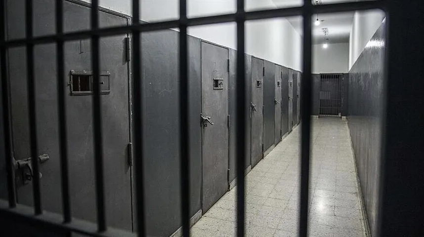 Tamni hodnik zatvora sa nizom metalnih vrata i rešetkama u prvom planu.