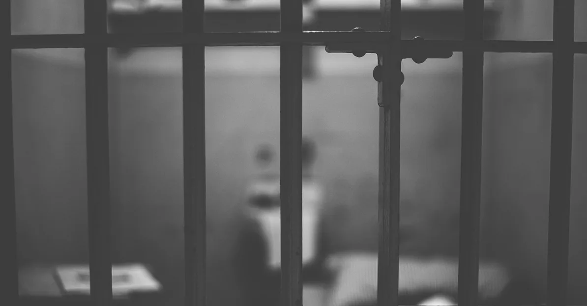 Crno-bijela fotografija zatvorske ćelije s debelim metalnim rešetkama u prvom planu. U pozadini, iza rešetaka, nazire se zamagljena figura osobe i krevet.