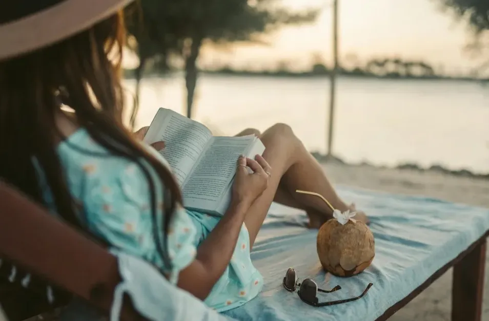 Žena u šeširu i ljetnoj haljini čita knjigu na ležaljci na plaži, pored nje je kokos sa slamkom i sunčane naočale.