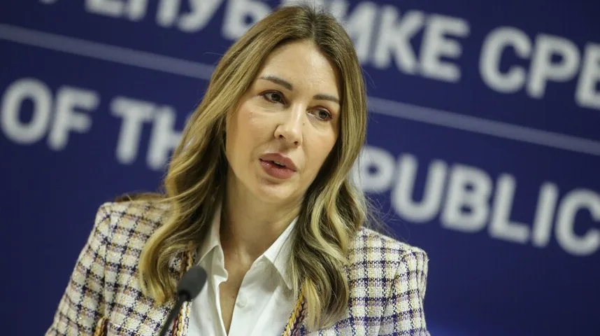 Žena u kariranom sakou govori ispred plave pozadine sa bijelim slovima