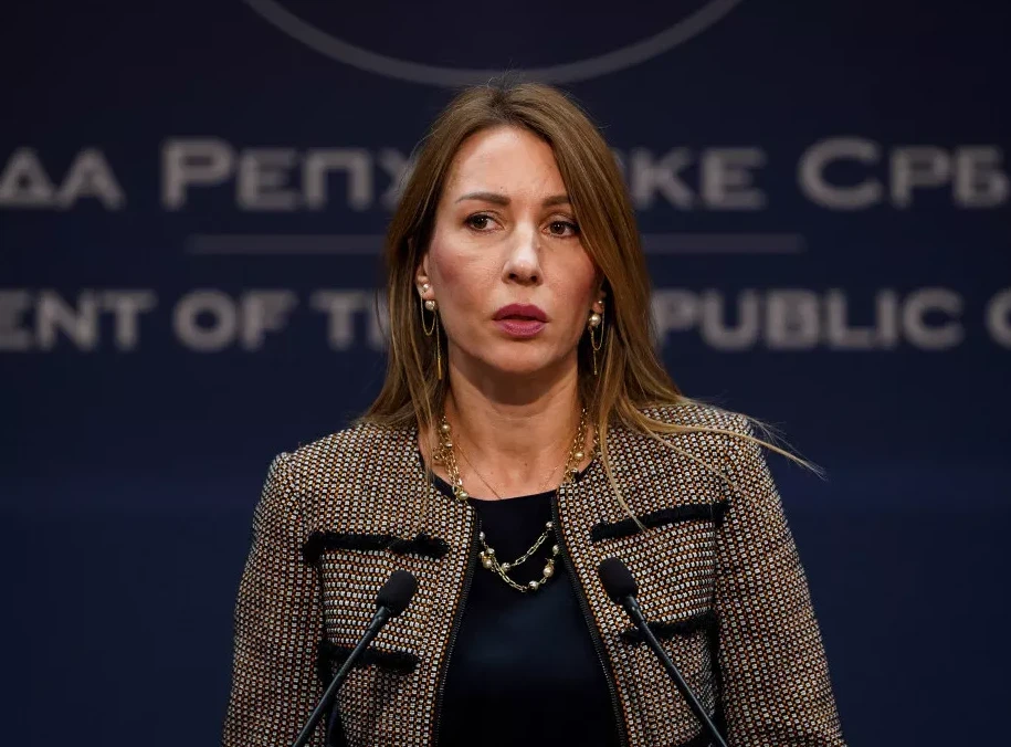 Žena sa smeđom kosom i naušnicama s biserima, obučena u kariranu jaknu, stoji ispred dva mikrofona na konferenciji za medije.
