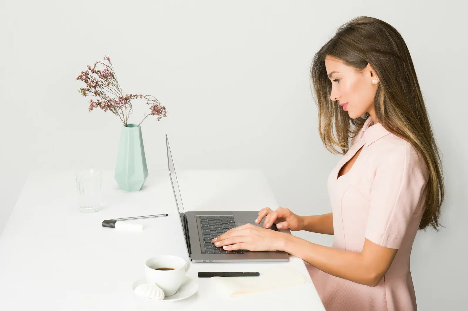 Mlada žena u roze haljini radi na svom laptopu za bijelim stolom, s kafom i cvijećem pored.