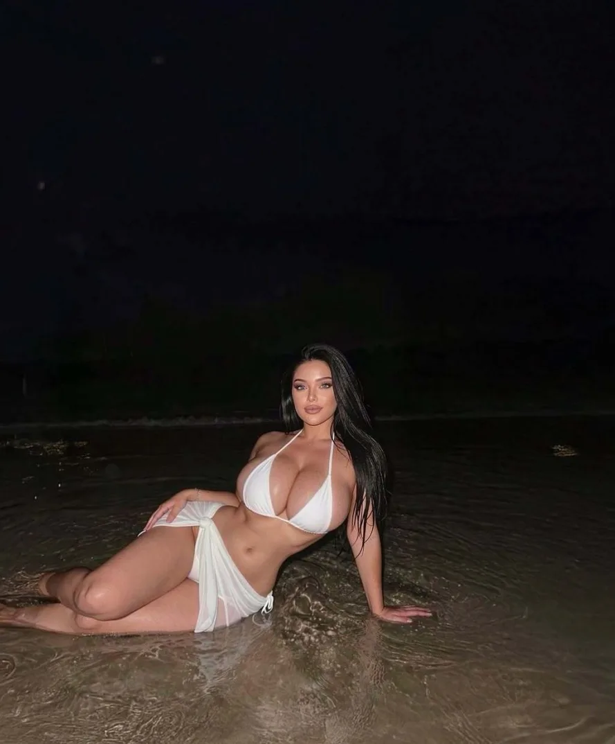 Žena u bijelom bikiniju sjedi u plićaku na plaži tokom noći.