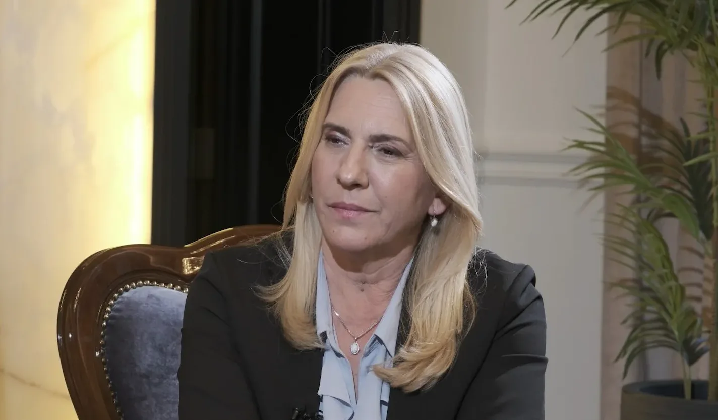 Žena sa plavom kosom, u crnom sakou i plavoj košulji, sjedi u fotelji.