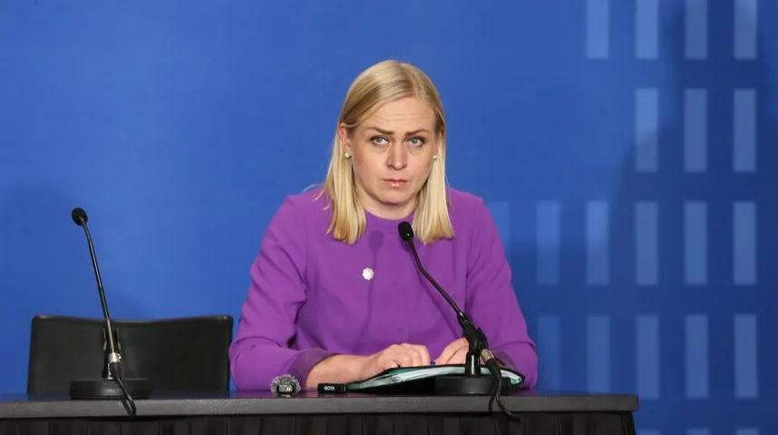 Žena sa plavom kosom u ljubičastoj haljini sjedi za stolom sa dva mikrofona i dokumentima.