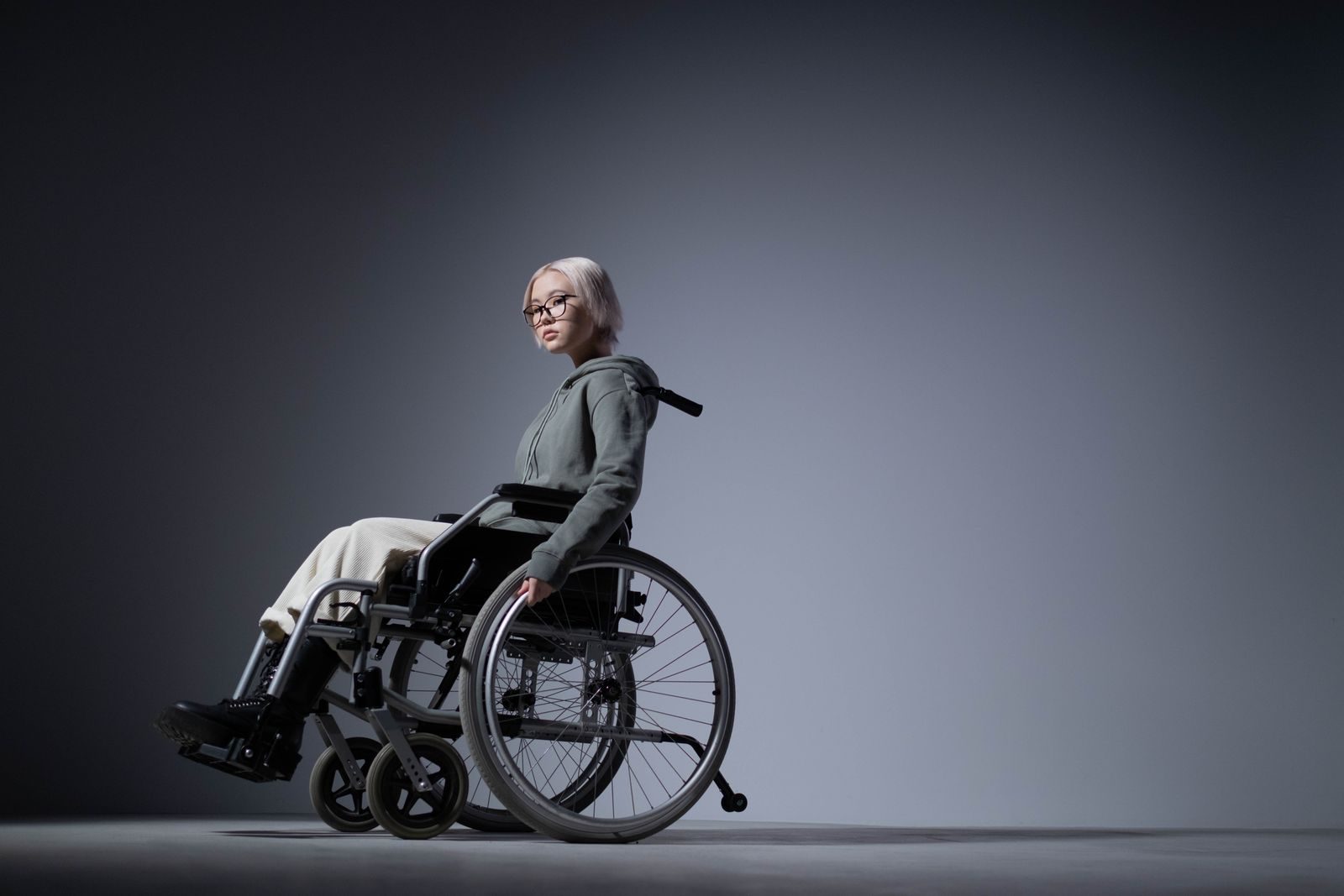 Studio portrait of an Asian woman in a wheelchair, wearing a gray hoodie and white pants.