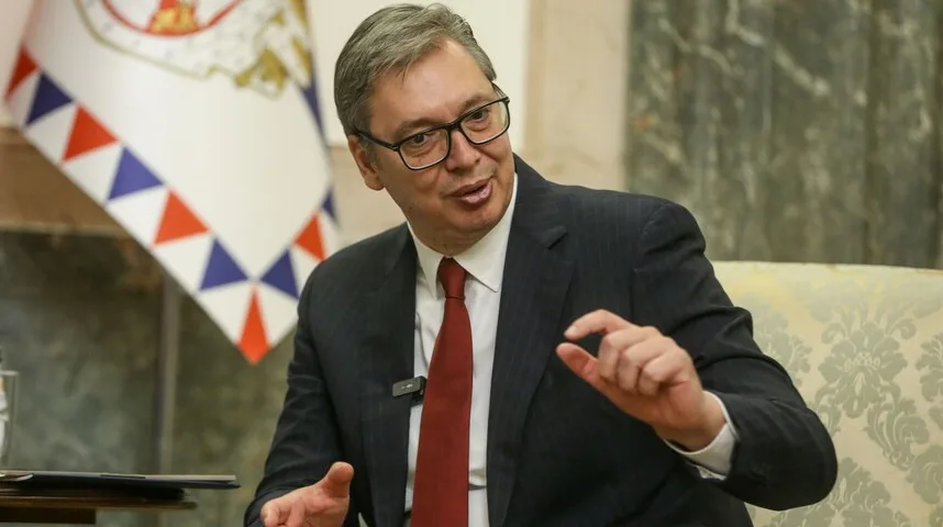 Aleksandar Vučić u tamnom odijelu, bijeloj košulji i crvenoj kravati, noseći naočare, drži lijevu ruku podignutu i gestikulira prstima desne ruke.