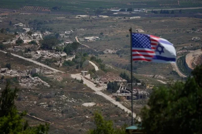 Američka i izraelska zastava istaknute u prvom planu sa pogledom na uništeno selo u pozadini.