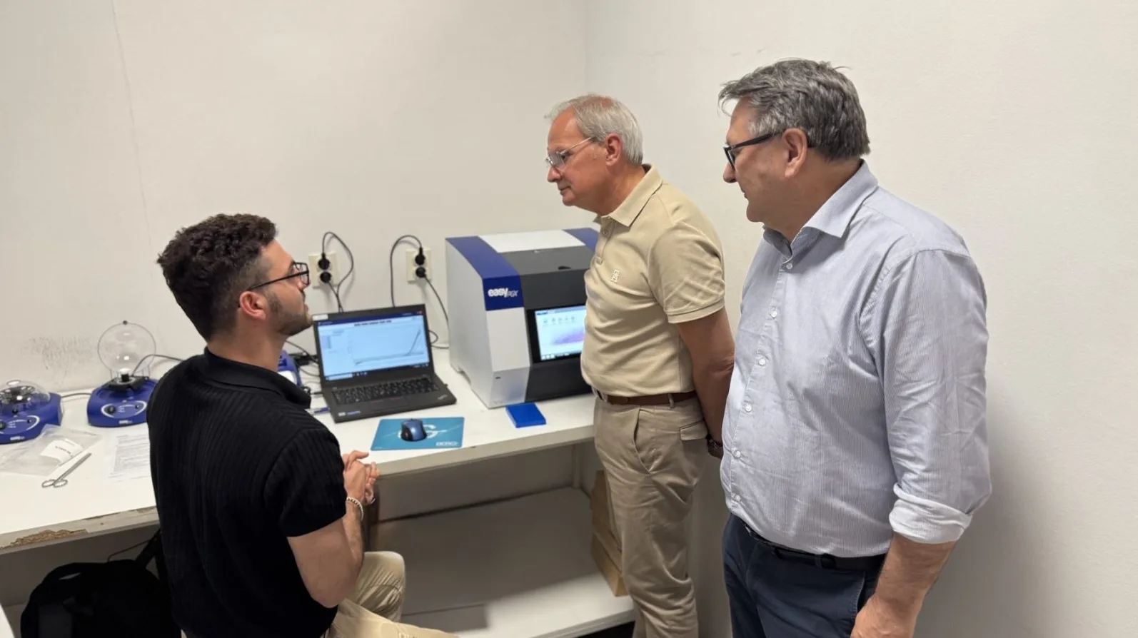 Tri osobe razgledaju modernu laboratorijsku opremu, uključujući PCR mašinu i laptop sa prikazom grafikona.