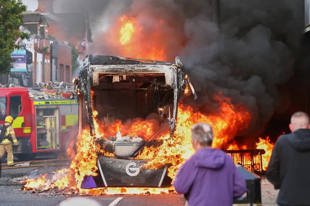 Autobus u plamenu sa vatrogasnim vozilom u pozadini i ljudima koji promatraju.