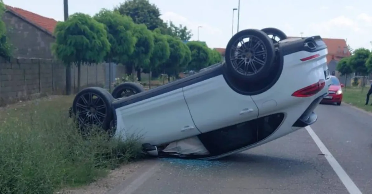 Bijeli automobil prevrnut na krovu na asfaltiranom putu, pored zelene trave i ograde od betonskih blokova. U pozadini se vide stabla, kuća s crvenim krovom, crveni automobil i osoba u uniformi.