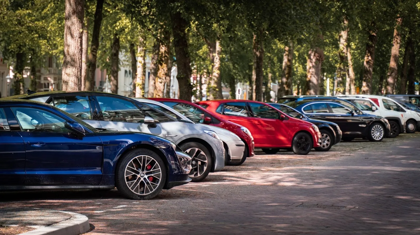 Više automobila različitih boja, uključujući tamnoplavi, srebrni i crveni, parkirani su u redu pored drvoreda na sunčan dan.