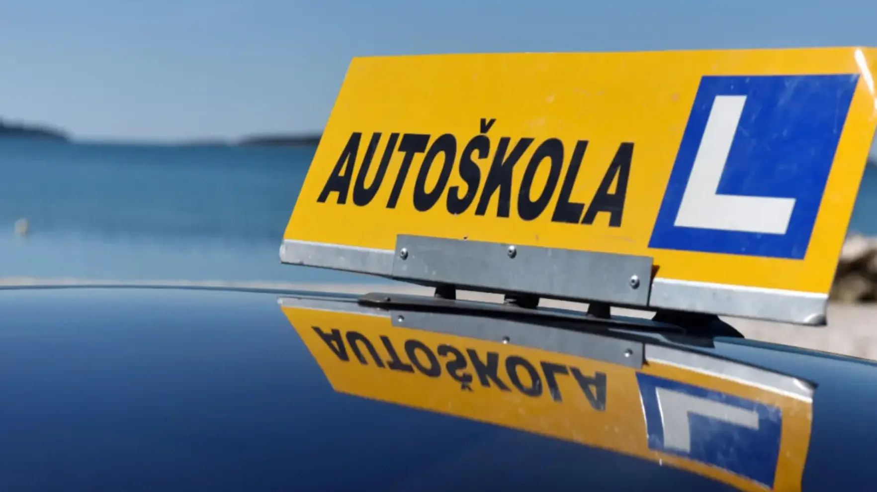 Žuti znak 'AUTOSKOLA' s plavim kvadratom i bijelim slovom 'L' na vrhu automobila, sa refleksijom znaka na tamnom krovu automobila.