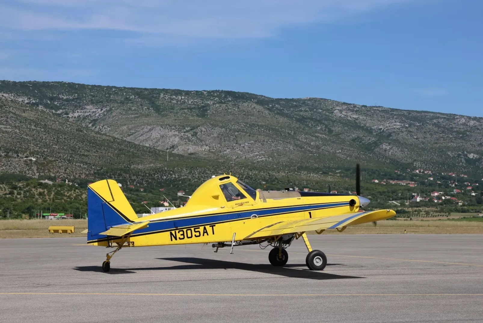 Žuti avion za prskanje sa plavim detaljima, registarske oznake N305AT, parkiran na aerodromskoj pisti sa brdima u pozadini.