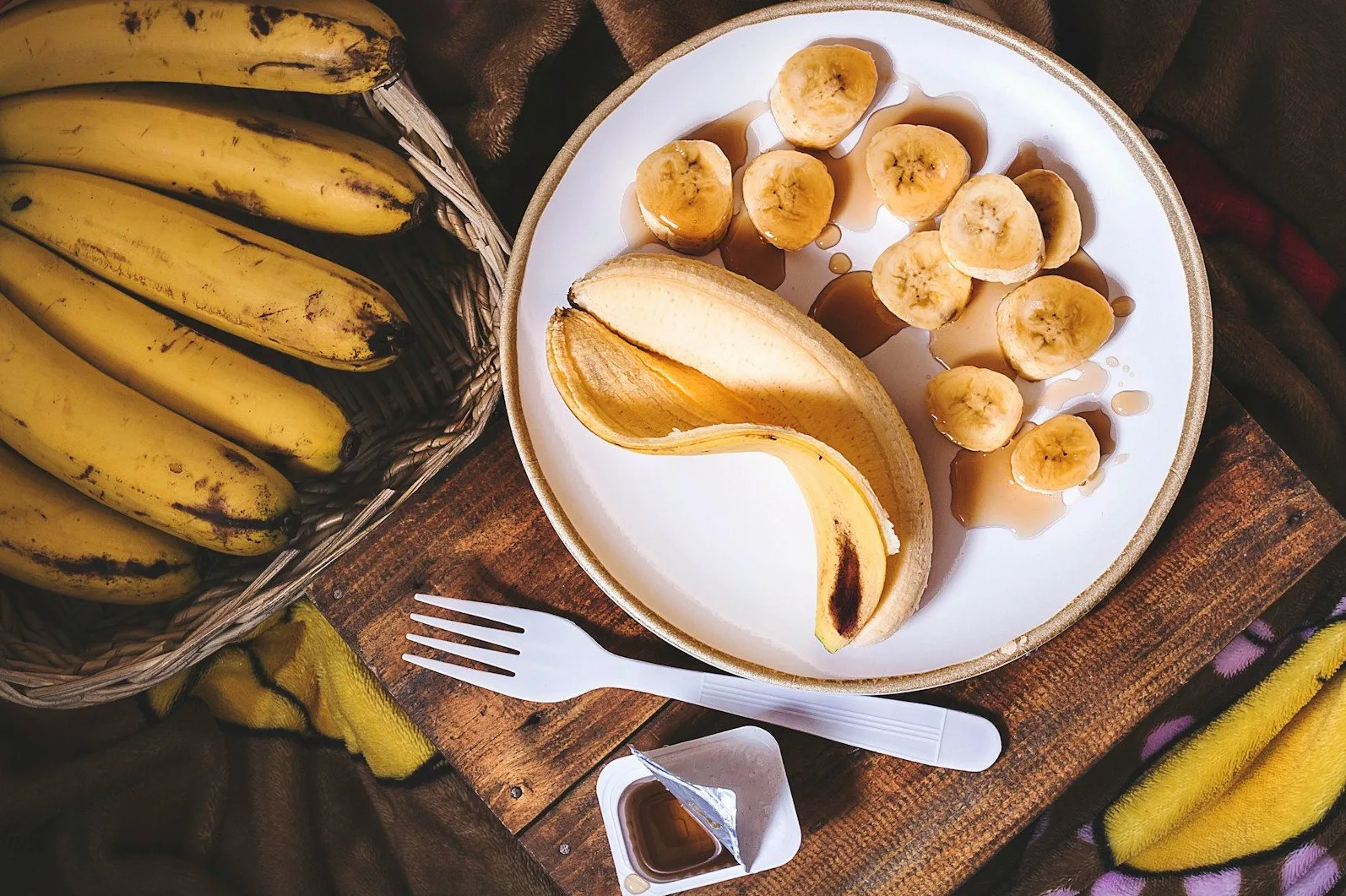 Blago oljuštena banana na tanjiru sa isječenim kriškama banane prelivenim medom, pored korpe punjene bananama, bijelog viljuškara i male kesice meda na drvenom stolu.