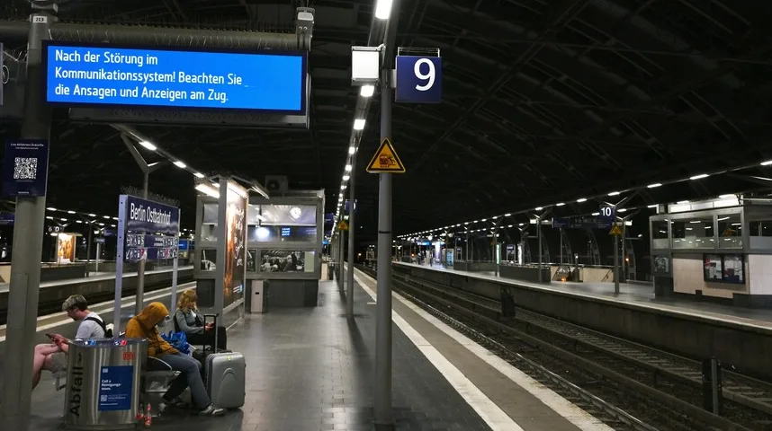 Putnici čekaju na peronu broj 9 na Berlinskoj stanici Ostbahnhof, sa plavim ekranom koji prikazuje obavještenje na njemačkom jeziku o kvaru komunikacijskog sistema.