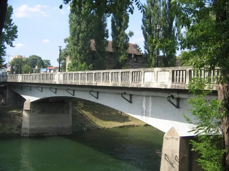 Betonski most sa ogradom od kamenih stubova i prečki, iznad zelene rijeke.