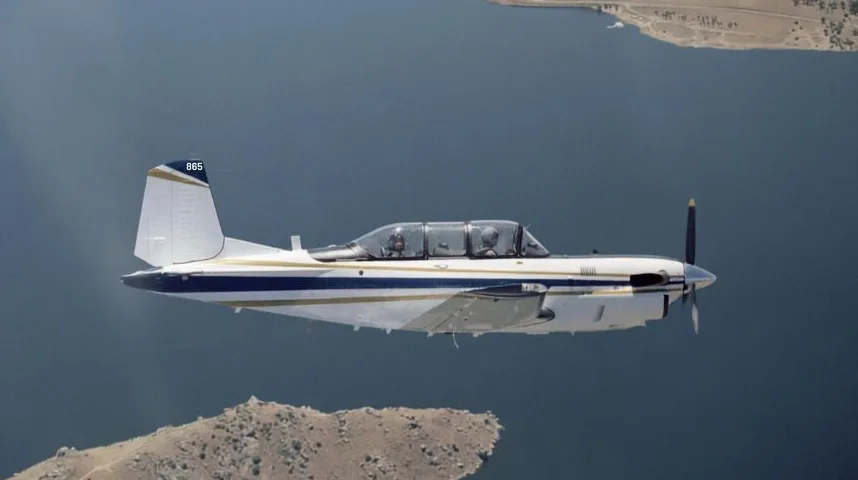 Bijeli mali avion sa plavom i zlatnom prugom leti iznad tamnom Plavog jezera ili mora, sa brdima u pozadini. Na repu aviona vidi se broj "865".
