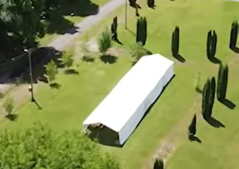 Bijeli šator postavljen na zelenom travnjaku pored staze i uređenih četinara.