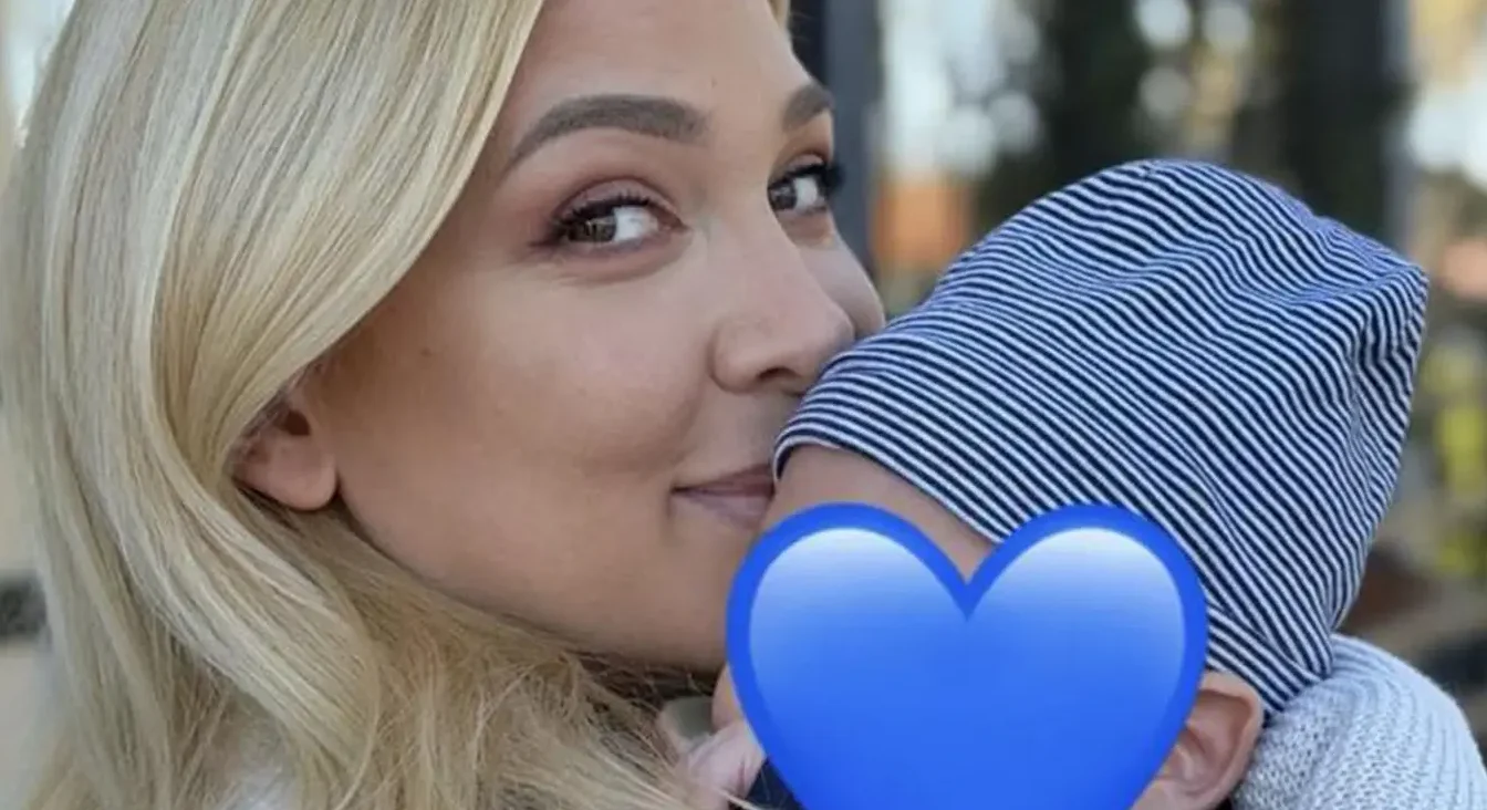 Blond majka s osmijehom na licu nježno grli svoju bebu, kojoj je lice prekriveno plavim srcem.