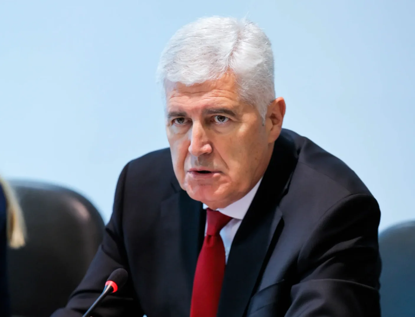 Fotografija prikazuje bosanskog političara sa sijedom kosom, obučenog u tamno odijelo, bijelu košulju i crvenu kravatu, kako sjedi za stolom i govori u mikrofon.