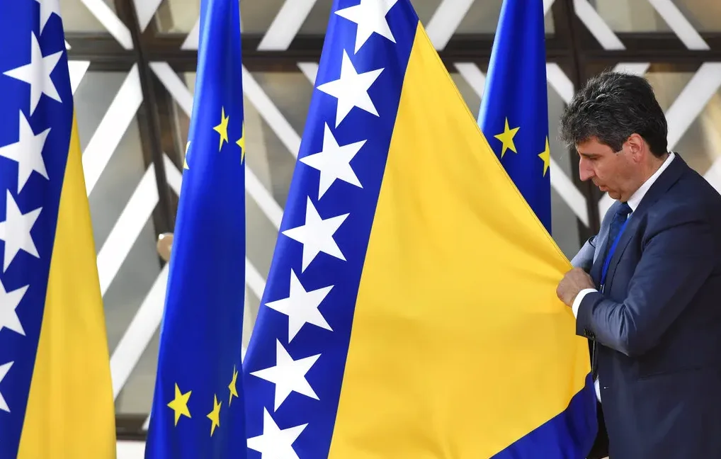 Muškarac u tamnom odijelu s plavom kravatom pažljivo drži zastavu Bosne i Hercegovine pored zastava Evropske unije.