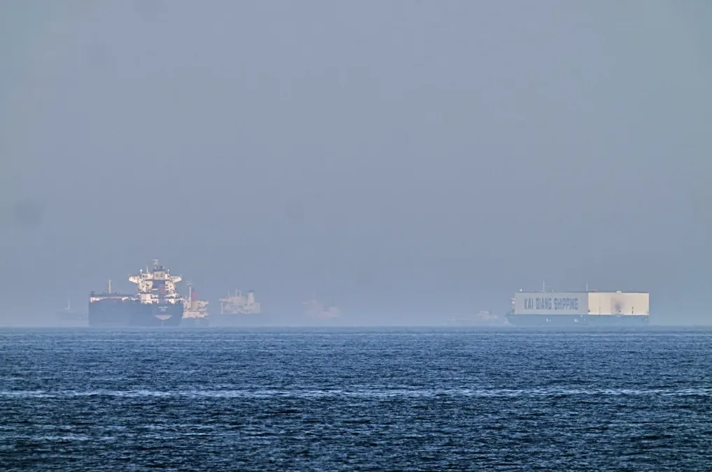 Veliki teretni brodovi usidreni na pučini, sa jednim brodom koji nosi natpis "KAI QIANG SHIPPING" u maglovitom danu.
