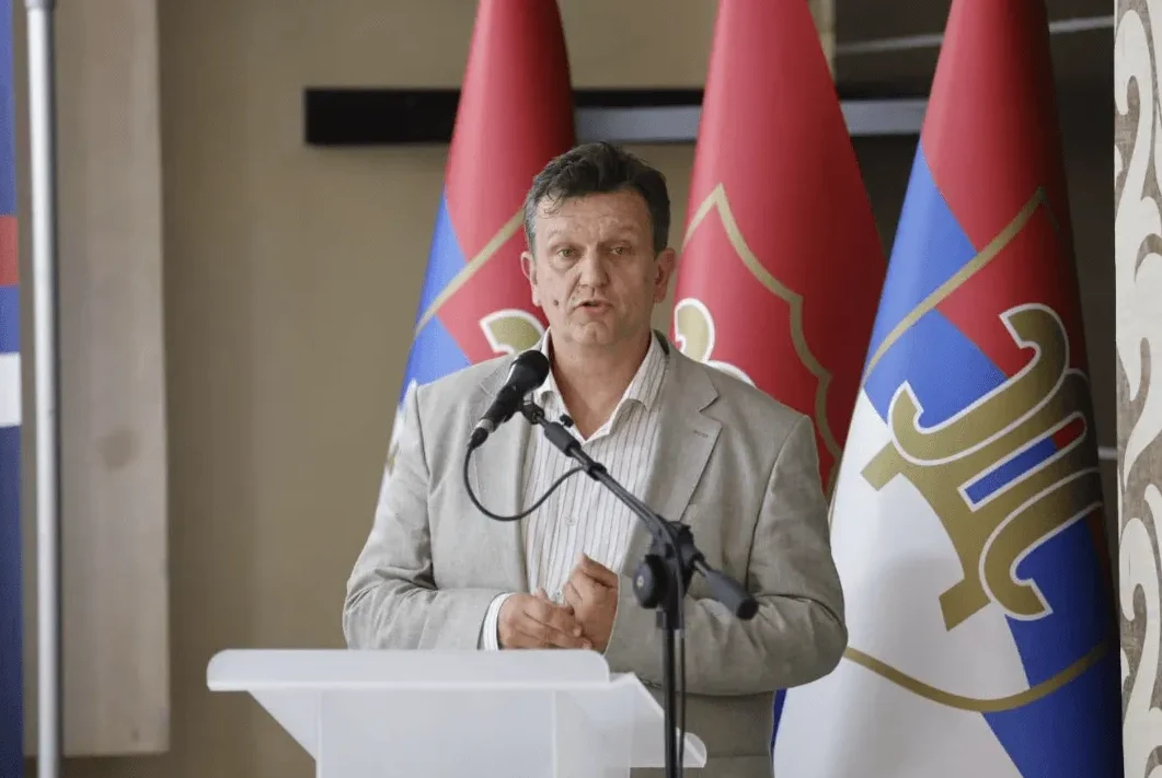 Muškarac u bijelom sakou i košulji stoji za govornicom ispred tri zastave.