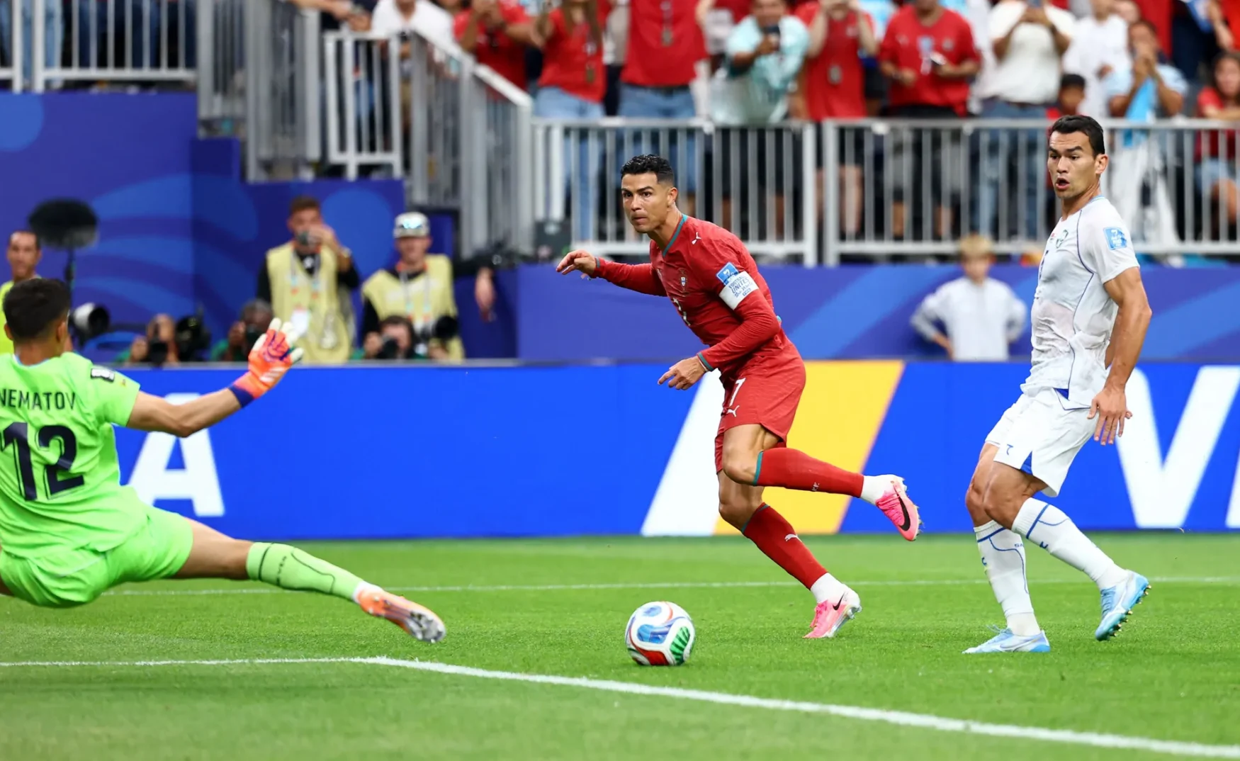 Fudbaler u crvenom dresu, za kojeg se pretpostavlja da je Kristijano Ronaldo, pokušava postići gol udarcem prema golu, dok golman u zelenom dresu pokušava odbraniti šut.