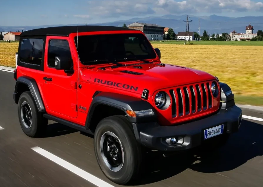 Crveni Jeep Wrangler Rubicon vozi se na asfaltiranom putu sa poljima i zgradama u pozadini.