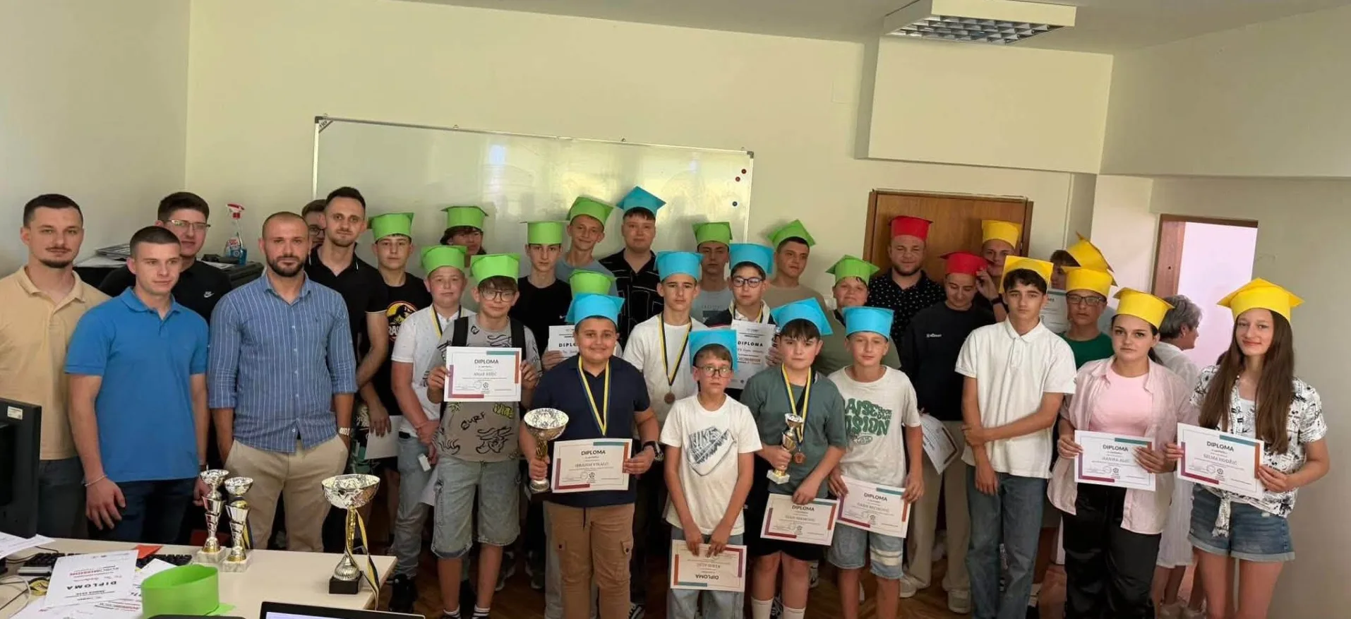 Grupa mladih ljudi, uglavnom dječaka i nekoliko djevojaka, pozira sa diplomama i trofejima, noseći šarene taboršake kape.
