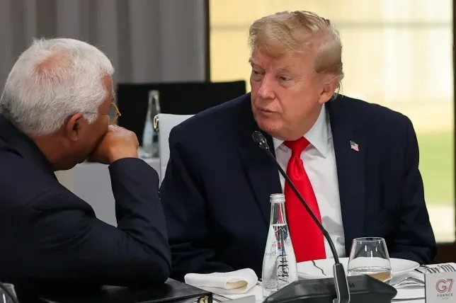Donald Tramp u tamnom odijelu i crvenoj kravati razgovara sa drugim muškarcem sijede kose koji sjedi lijevo od njega i naslonio je ruku na obraz.