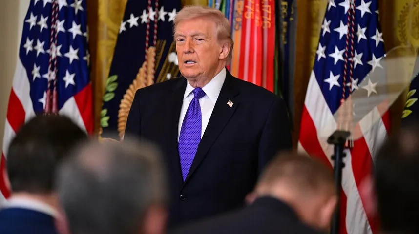 Donald Tramp u crnom odijelu i ljubičastoj kravati, stojeći ispred američke zastave i nekoliko drugih zastava.